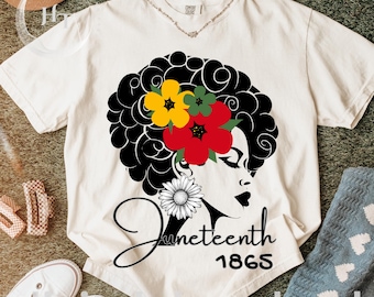 May include: Cream-colored t-shirt featuring a graphic of a woman's profile with a floral afro and a daisy earring. The text "Juneteenth 1865" is written in a script font. The shirt is displayed on a green surface.