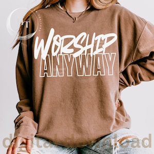 May include: A brown sweatshirt with the words "WORSHIP ANYWAY" in white block letters. The sweatshirt has long sleeves and a crew neck. The text is centered on the front of the shirt.