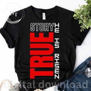 May include: A black short-sleeve t-shirt featuring the white text "STORY" and "HE IS RISEN" alongside the large, bold red vertical text "TRUE". The shirt has a crew neckline and is displayed flat with a visible brand label at the inner collar.