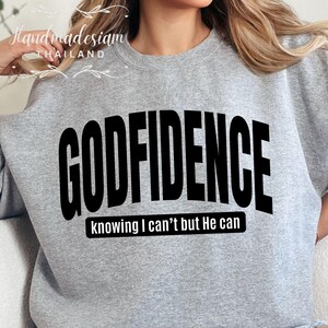 May include: Gray sweatshirt with the text "GODFIDENCE" in black, arched across the front. Below the text is the phrase "knowing I can't but He can" in smaller black text.