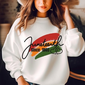 以下が含まれることがあります： 虹色のデザインと「Juneteenth Since 1865」の文字、そして2つの絡み合ったハートが描かれた白いスウェットシャツです。