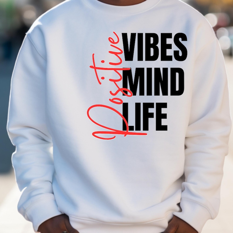 Op de afbeelding: Witte sweatshirt met de woorden "Positive Vibes Mind Life" in zwarte en rode letters. Het woord "Positive" is verticaal in rood schrift aan de linkerkant geschreven.