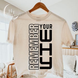 May include: A cream-coloured short-sleeve t-shirt featuring bold black vertical text on the front that reads REMEMBER YOUR WHY. The shirt has a crew neckline and is displayed on a wooden hanger, showcasing a simple and clean graphic design.