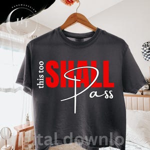 May include: Dark grey t-shirt with the words "this too SHALL pass" in red and white. The text is centered on the shirt. The shirt is hanging on a wooden hanger.