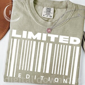 Könnte beinhalten: Ein salbeigrünes T-Shirt mit dem weißen Schriftzug "LIMITED EDITION" und einem Barcode-Design. Eine silberne Halskette mit rosa Anhänger liegt auf dem Shirt. Das Shirt hat ein Comfort Colors-Etikett.
