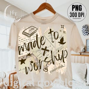 May include: Cream-colored t-shirt with the words "made to worship" in a handwritten style. The design includes a cross, a dove, stars, and a book. The text "psalm 95:1" is also visible.