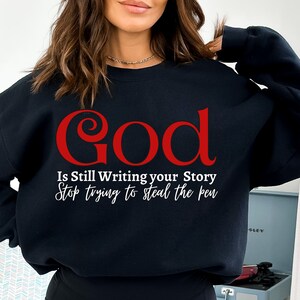 Op de afbeelding: Zwart sweatshirt met de tekst "God Is Still Writing Your Story Stop Trying To Steal The Pen" in witte en rode letters.
