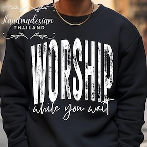 May include: Black sweatshirt with the word "WORSHIP" in large, distressed white letters across the front. Below the word "WORSHIP" is the phrase "while you wait" in a cursive font. The sweatshirt has a crew neck.