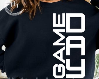May include: A black long-sleeve sweatshirt featuring the white text GAME DAY printed vertically in a bold, sans-serif font on the right side. The garment has a crew neckline and a solid black fabric construction throughout the front panel.