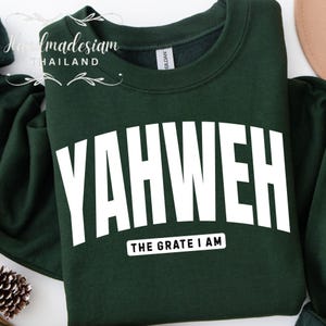 May include: A dark green sweatshirt with the word "YAHWEH" in large, bold, white letters across the front. Below the word "YAHWEH" is the text "THE GRATE I AM" in a smaller, white font. The sweatshirt is a solid color.