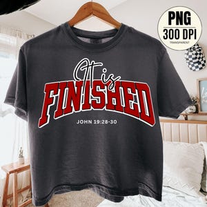 May include: Dark gray t-shirt with the words "It is FINISHED" in red outlined in white. The text "JOHN 19:28-30" is below the main text. The shirt is hanging on a wooden hanger.