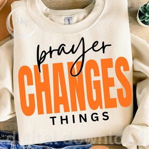 May include: A cream-coloured crewneck sweatshirt featuring the black cursive text "prayer" above the large, bold, orange block-letter text "CHANGES" and the black capitalised text "THINGS" centred on the front of the garment.