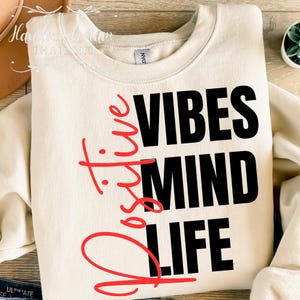 Op de afbeelding: Cr&egrave;mekleurige sweatshirt met de tekst "Positive Vibes Mind Life" in zwarte en rode letters. De tekst is verticaal en horizontaal geplaatst. De sweatshirt heeft een ronde hals.