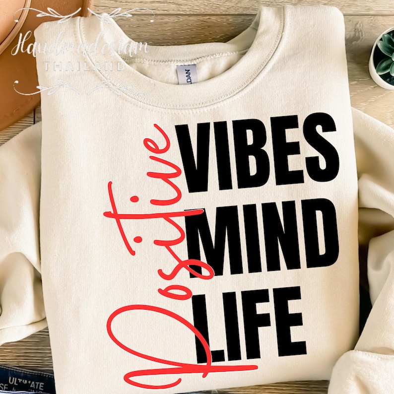 Op de afbeelding: Cr&egrave;mekleurige sweatshirt met de tekst "Positive Vibes Mind Life" in zwarte en rode letters. De tekst is verticaal en horizontaal geplaatst. De sweatshirt heeft een ronde hals.