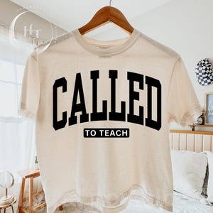Peut inclure: Un t-shirt à manches courtes de couleur crème avec le texte noir CALLED en grande police universitaire arquée, et les mots TO TEACH imprimés dans un petit rectangle noir directement sous le texte principal sur le devant.
