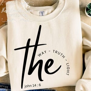 Puede incluir: Sudadera color crema con la palabra "The" en letras negras grandes y en negrita. Las palabras "WAY - TRUTH - LIGHT" están en semicírculo alrededor de la palabra "The". El texto "John 14:6" está en la parte inferior.