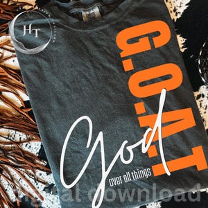 May include: A dark charcoal grey t-shirt featuring the vertical orange text G.O.A.T. and the white cursive script text God over all things. The shirt has a crew neckline and a Comfort Colors brand label visible at the inner collar area.