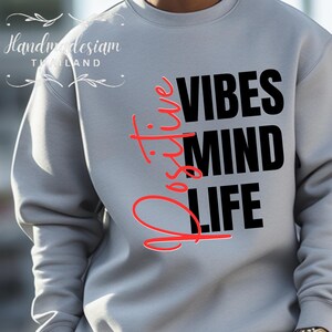 Op de afbeelding: Een lichtgrijs sweatshirt met de woorden "Positive Vibes Mind Life" in zwarte en rode letters. Het woord "Positive" is verticaal geschreven in rood cursief. De sweatshirt heeft lange mouwen.