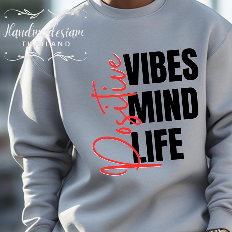 Op de afbeelding: Een lichtgrijs sweatshirt met de woorden "Positive Vibes Mind Life" in zwarte en rode letters. Het woord "Positive" is verticaal geschreven in rood cursief. De sweatshirt heeft lange mouwen.