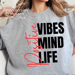 Op de afbeelding: Een grijze sweatshirt met de woorden "Positive Vibes Mind Life" in zwarte en rode letters. Het woord "Positive" is geschreven in een cursief lettertype. Het merk "Handmadesiam Thailand" staat in de linkerbovenhoek.