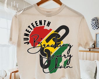 May include: A cream-colored t-shirt with the words "Juneteenth Breaking Every Chain" in black script. The design includes a red, yellow, and green graphic. The shirt is hanging on a wooden hanger.