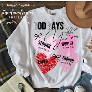 May include: White sweatshirt with a graphic design of three hearts with inspirational messages and bible verses. The text reads "God Says You Are Strong, Worthy, Enough".