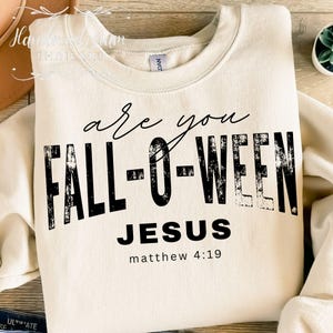 Puede incluir: Sudadera color crema con el texto "are you FALL-O-WEEN JESUS" en negro, con "matthew 4:19" debajo. La sudadera es de cuello redondo.
