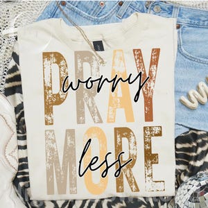 May include: A cream-colored short-sleeve t-shirt featuring the distressed block-letter text PRAY MORE in shades of brown, tan, and beige, overlaid with the black cursive script text worry less. The shirt is displayed with denim and accessories.