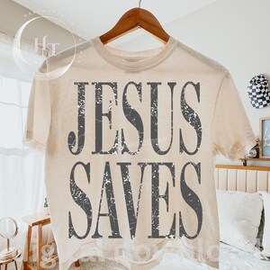 May include: Cream-colored t-shirt with the text "JESUS SAVES" in distressed gray lettering. The shirt is hanging on a wooden hanger. The overall theme is religious and inspirational.