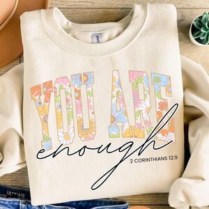 May include: Cream-coloured sweatshirt with the words "YOU ARE enough" in a floral design. The text "2 CORINTHIANS 12:9" is below the word "enough". The sweatshirt is folded on a wooden surface.