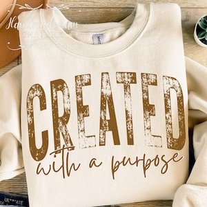 Puede incluir: Sudadera color crema con las palabras "CREATED with a purpose" impresas en marrón. La sudadera está doblada, con el texto visible. El diseño tiene un aspecto envejecido y vintage.