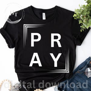 Może przedstawiać: Czarny t-shirt z krótkim rękawem z napisem PRAY zapisanym dużymi białymi wielkimi literami. Tekst jest zamknięty w białej ramce graficznej składającej się z linii narożnych. Koszulka ma okrągły dekolt i standardowy krój z podwiniętymi rękawami.