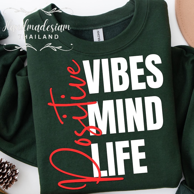 Op de afbeelding: Een donkergroene sweatshirt met de woorden "Positive Vibes Mind Life" in witte en rode letters. De sweatshirt heeft een ronde hals en lange mouwen. De merknaam "GILDAN" is zichtbaar op het label.
