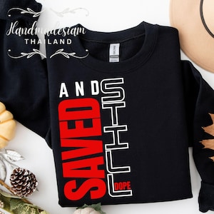 May include: Black sweatshirt with white text that reads "Still Dope" and "Saved" in red. The text is vertically aligned on the left side of the sweatshirt.