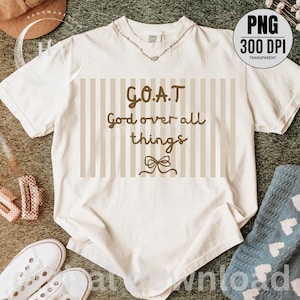 May include: Cream-colored t-shirt with the text "G.O.A.T God over all things" in brown script. The text is set against a beige striped background with a bow graphic. The shirt has a V-neck and is styled with a necklace.