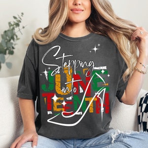 May include: A black t-shirt with a distressed graphic design that says "Stepping into Juneteenth" in white text with a red, yellow, and green color scheme. The graphic design includes a stylized high heel shoe.