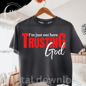 May include: Dark gray t-shirt with the text "I'm just out here TRUSTING God" in red and white. The shirt is hanging on a wooden hanger. The text is centered on the front of the shirt.
