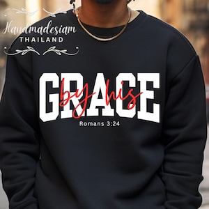 Puede incluir: Sudadera negra con las palabras "GRACE by his" en letras blancas y escritura cursiva roja. El texto "Romans 3:24" está debajo. La sudadera tiene cuello redondo.