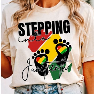May include: A white t-shirt with the text "Stepping into Juneteenth" in black lettering. The text is surrounded by a red, yellow, and green outline of an African continent with two black footprints. Each footprint has a heart-shaped design with the colors of the Pan-African flag.