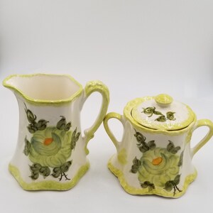 May include: A cream-colored ceramic creamer and sugar bowl set. The set features a floral design with green and orange accents. The rims and handles are painted in a light green color.