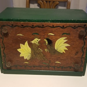 May include: A wooden bread box with a green lid and a brown base. The base has a painted design of two yellow and brown roosters with a yellow background.