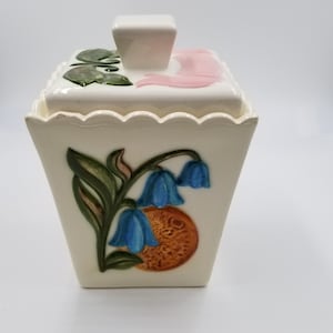 May include: Cream-colored ceramic container with a square lid and a scalloped edge. The container features a raised design of bluebell flowers and an orange-brown fruit. The lid has a white square handle and pink and green floral accents.