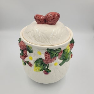 Peut inclure: Un pot en céramique blanc avec un couvercle, orné de feuilles en relief et de fraises en 3D. Le couvercle comporte deux poignées en forme de fraise. Le pot est décoré de fraises, de fleurs blanches et de feuilles vertes.