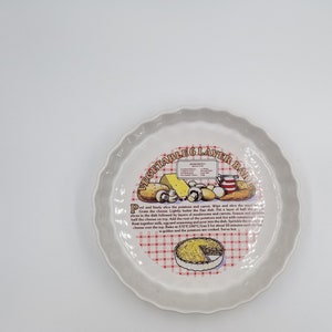 May include: A white ceramic baking dish with a red and white checked pattern and the text "VEGETABLE 6 LAYER BAKE" printed on it. The dish features a colourful illustration of ingredients for a vegetable bake, including potatoes, carrots, mushrooms, cheese, and onions.