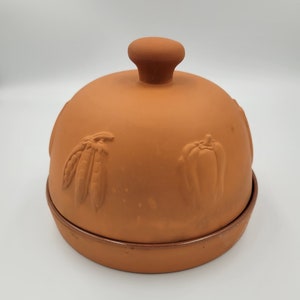 May include: A terracotta cloche with a round base and a domed lid. The lid features embossed vegetable designs, including peas and a bell pepper. A small knob is on top. The cloche is designed for cooking and serving.