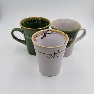 May include: Three ceramic mugs in shades of green and off-white. The mugs have handles and decorative rims. One mug is a tumbler shape with a black squiggle design. The mugs are on a white background.