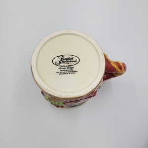 Pode incluir: Uma caneca de cer&acirc;mica com um design colorido, com uma al&ccedil;a vermelha e um padr&atilde;o detalhado ao redor da borda. A parte inferior da caneca exibe o texto "Certified International" e "Made in China". A caneca &eacute; projetada por Susan Winget.