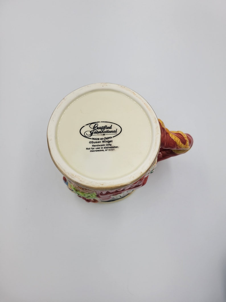 Pode incluir: Uma caneca de cer&acirc;mica com um design colorido, com uma al&ccedil;a vermelha e um padr&atilde;o detalhado ao redor da borda. A parte inferior da caneca exibe o texto "Certified International" e "Made in China". A caneca &eacute; projetada por Susan Winget.