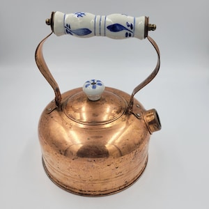 May include: A copper teapot with a white and blue porcelain handle and lid knob. The teapot has a shiny copper finish and a rounded body.
