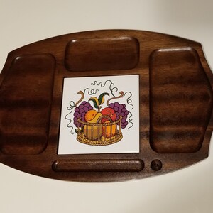 May include: A wooden serving tray with a decorative tile insert featuring a basket of fruit. The tray has a dark brown finish and includes three recessed areas for holding items. The tile insert displays grapes, apples, and pears.